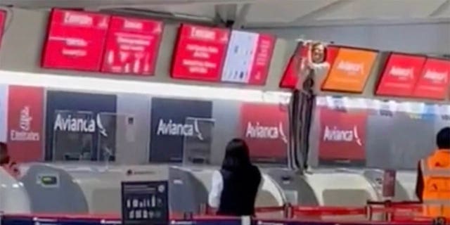 Andere passagiers op de luchthaven kunnen de uit de hand gelopen persoon - die bij de incheckbalie staat en er een scherm boven houdt - van ver zien.